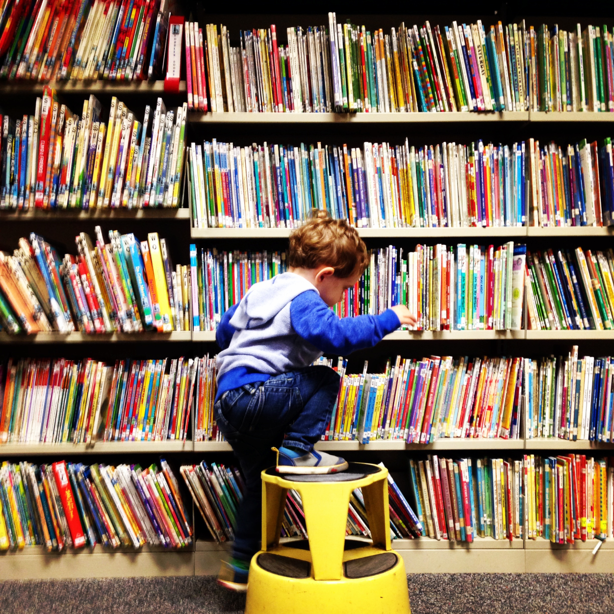 Img Block2 Toddler on a yellow stool in the Children's room Toddler and Junior reader section. There are multiple shelves and books in the background.